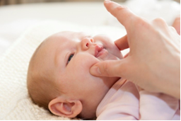 Photo of an adult touching a baby's face with more fingers. The ring and little fingers are placed on the front part of the floor of the mouth, under the chin. The thumb and middle finger are placed on each cheek. The index finger is ready to be inserted into the mouth.