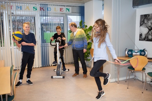 En pige danser i forgrunden, imens en dreng står med en bold og en anden dreng cykler på motionscykel i baggrunden.