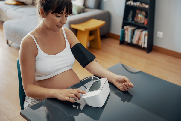 Foto af gravid kvinde, der sidder afslappet p&aring; opretst&aring;ende stol foran bord. Kvinden har manchet til m&aring;ling af blodtryk om overarmen og blodtryksapparatet p&aring; bordet foran sig. Underarm og h&aring;nd hviler p&aring; bordet.